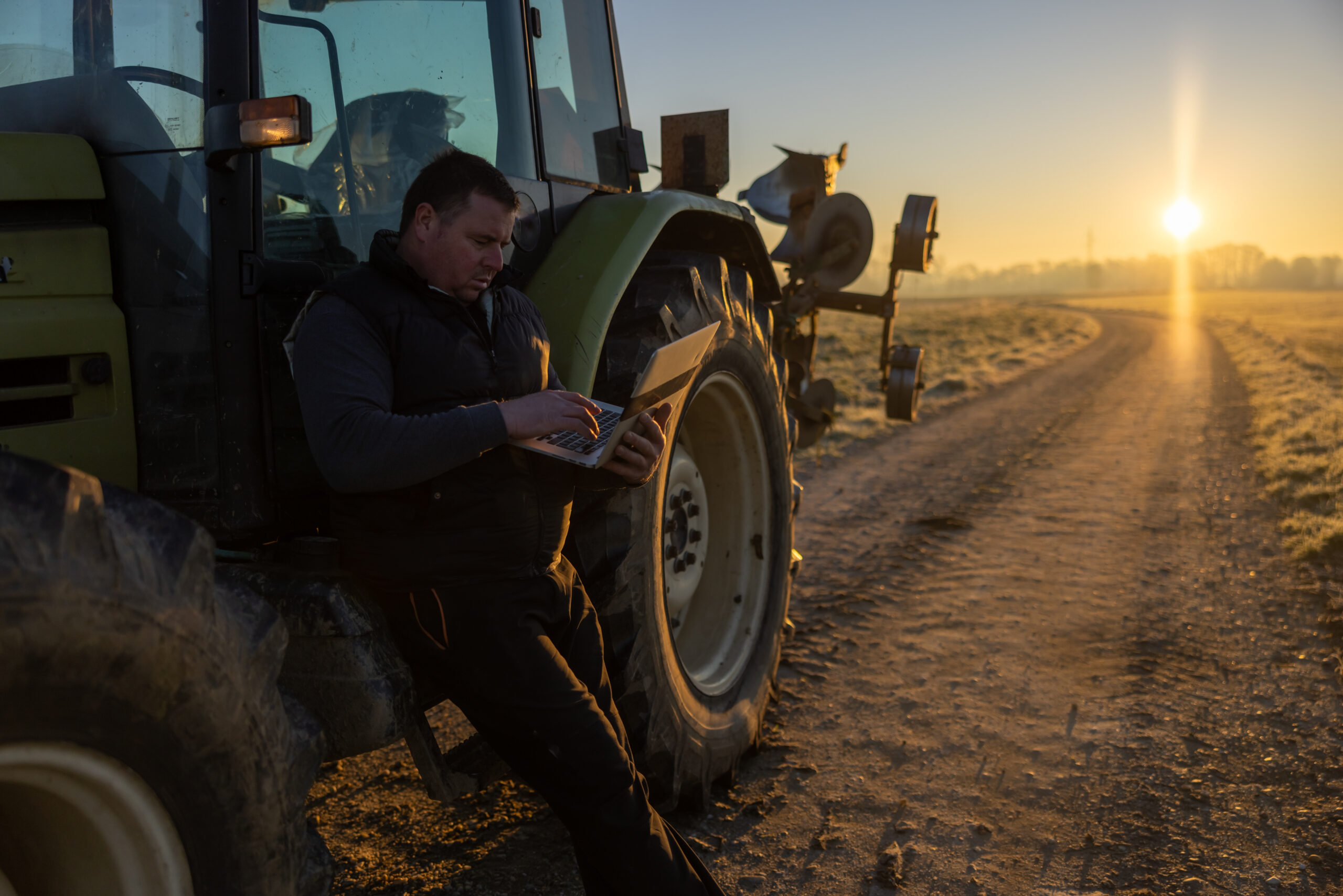 Man with short brown hair,wearing trousers,a gray pullover and black vest is using his laptop,concentrated facial expression,leaning against his green tractor in the evening parked on an agricultural path during sunset,side view